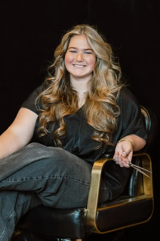 Smiling person with long, wavy hair sits in a chair, holding hairstyling scissors in one hand.