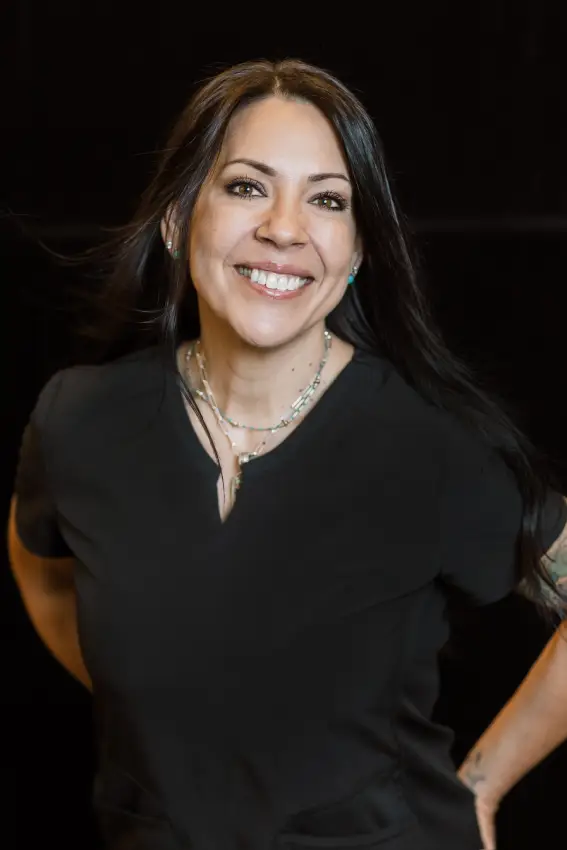Smiling woman with long black hair wearing a black top, standing with hands on hips.