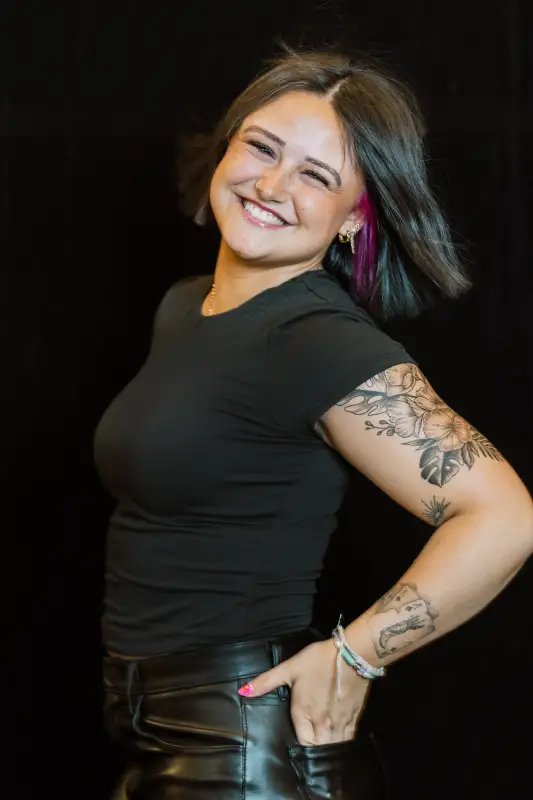 Smiling person with tattoos on arms, wearing a black shirt and leather pants, with a streak of pink in their hair.