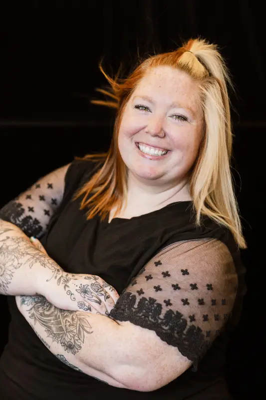 A person with long hair and tattoos on their forearms smiles, wearing a black top with lace sleeves.