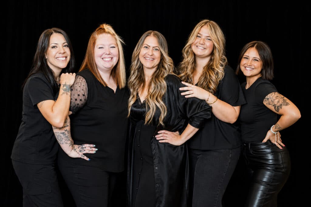 Five smiling women wearing black outfits stand together against a dark background.